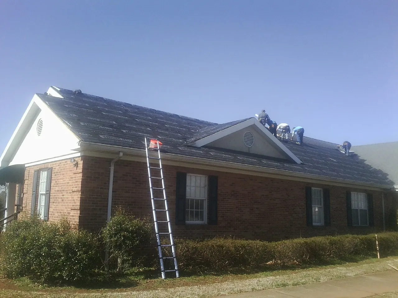 Crew tearing shingles off a brick ranch roof with a ladder set — Metal Roof Repair work by our crew in Bunnell
