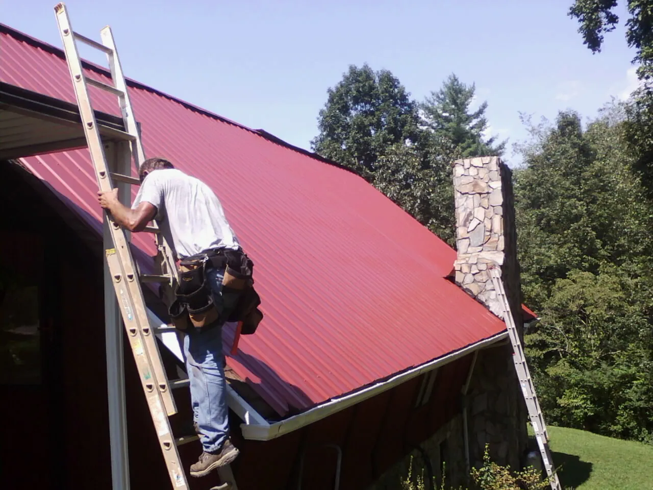Roofer on a ladder beside a red metal roof and stone chimney — Soffit & Fascia Repair by Roofing Brothers in Bunnell